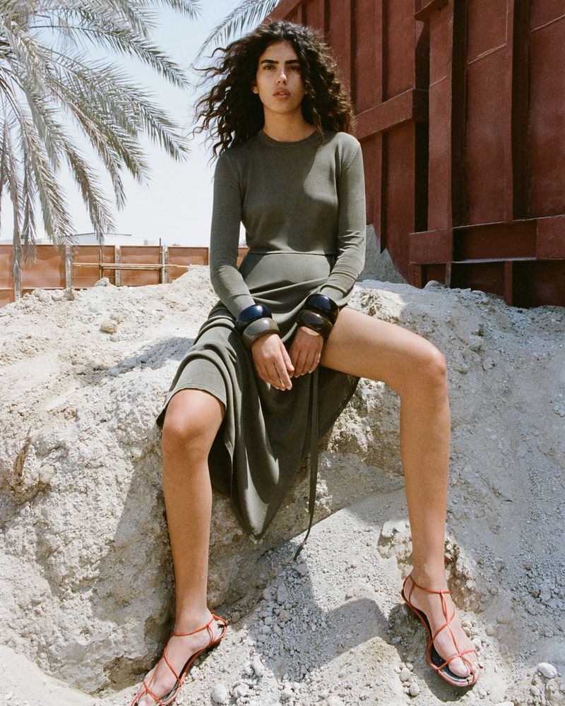 A woman with dark, curly hair sits on a rock pile wearing a long-sleeve olive-green dress and strappy sandals against an outdoor desert backdrop.