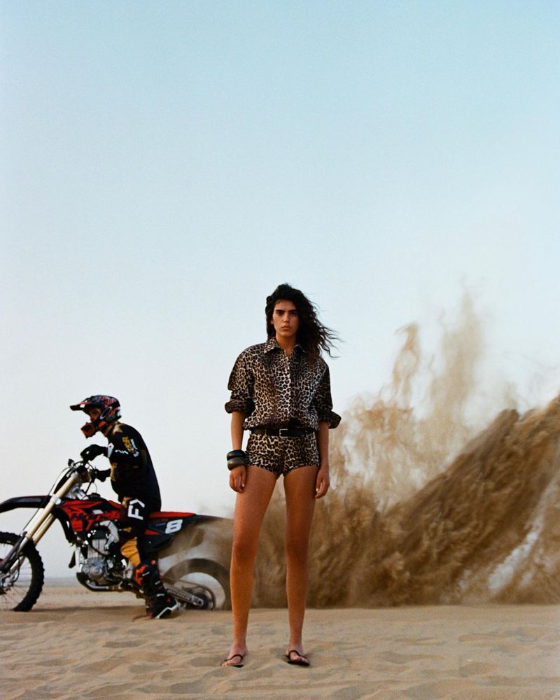 Woman in leopard print outfit stands in desert with motorcycle rider and dust cloud in the background under a light blue sky.