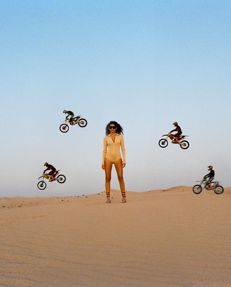 A woman stands in the desert with four motocross riders jumping in the air around her in a striking and surreal composition.