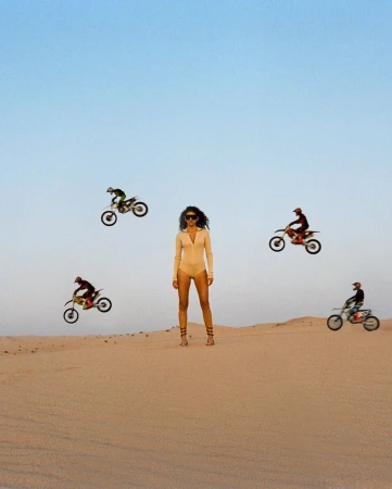 A woman stands in the desert with four motocross riders jumping in the air around her in a striking and surreal composition.