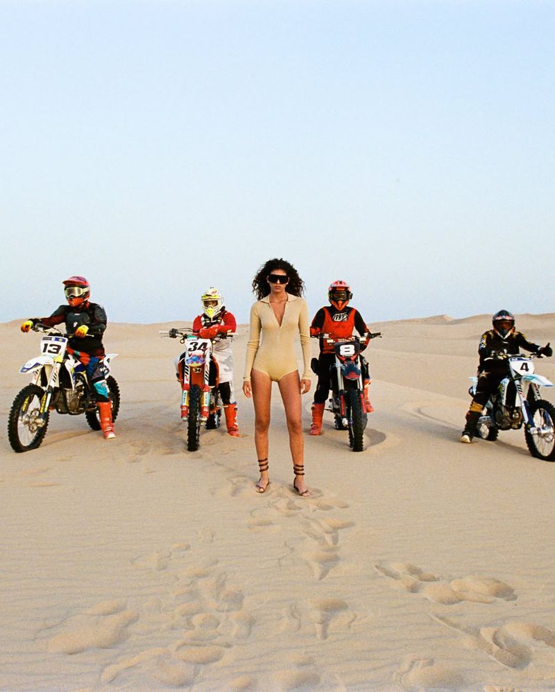 A group of people on dirt bikes posing on sand dunes with a woman in a light tan bodysuit standing in the center.