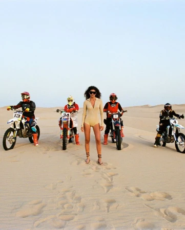 A group of people on dirt bikes posing on sand dunes with a woman in a light tan bodysuit standing in the center.