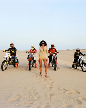 Model standing in desert sand wearing a beige bodysuit flanked by four motocross riders on dirt bikes