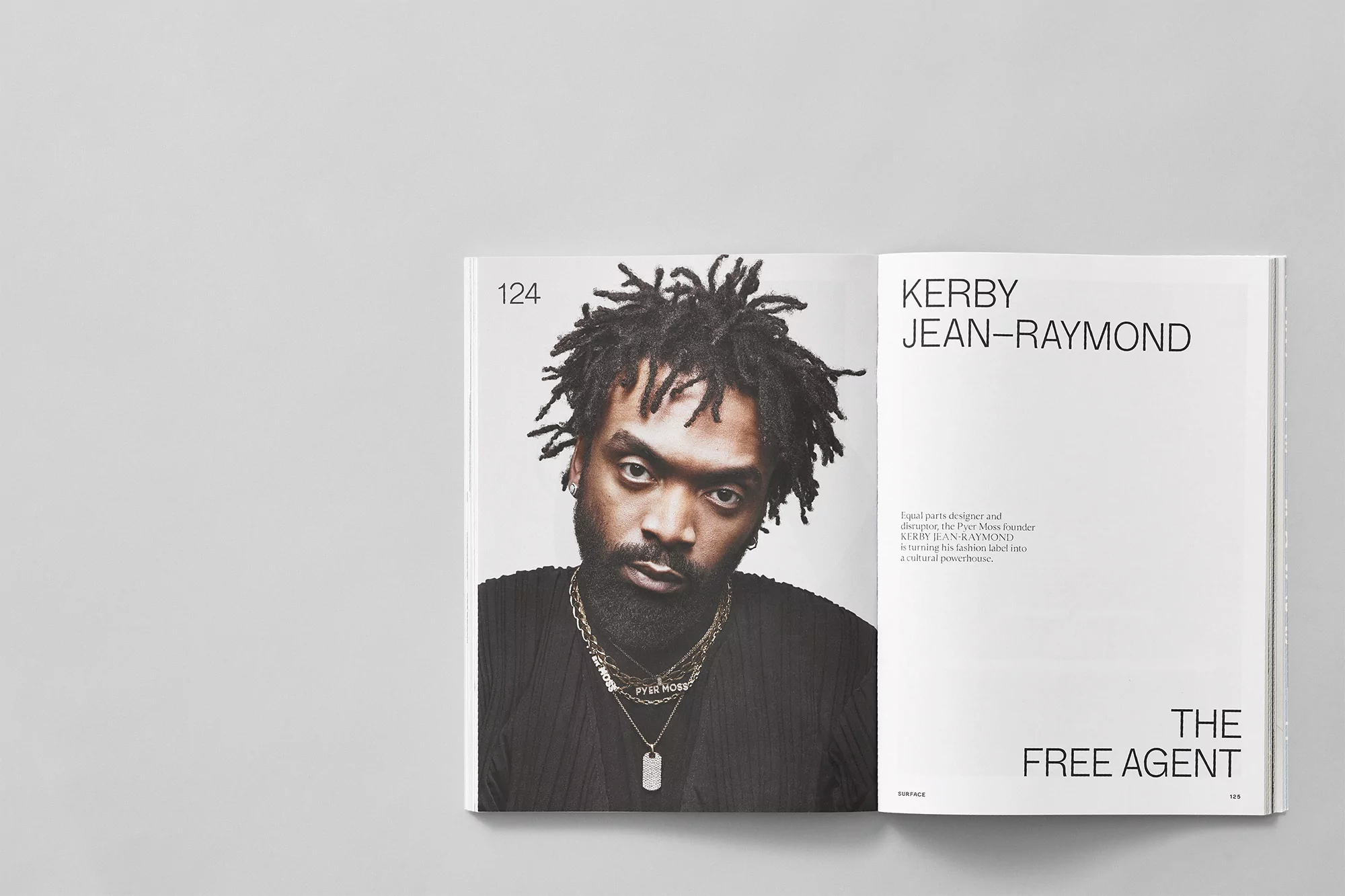 Portrait of Kerby Jean-Raymond in a magazine, showcasing his dark hair, beard, chains, and black shirt against a white backdrop.