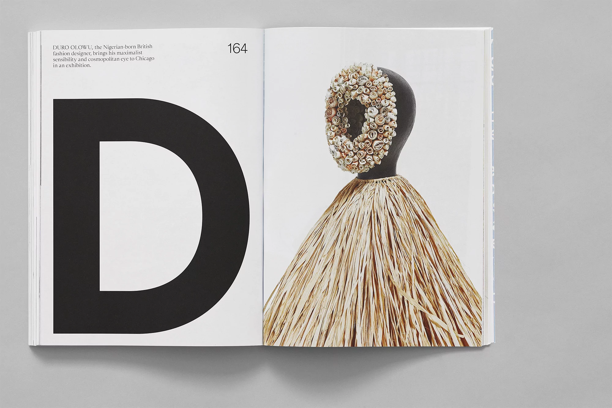 Open book featuring a bold black letter D and a mannequin head adorned with shells, paired with a raffia skirt-like garment.