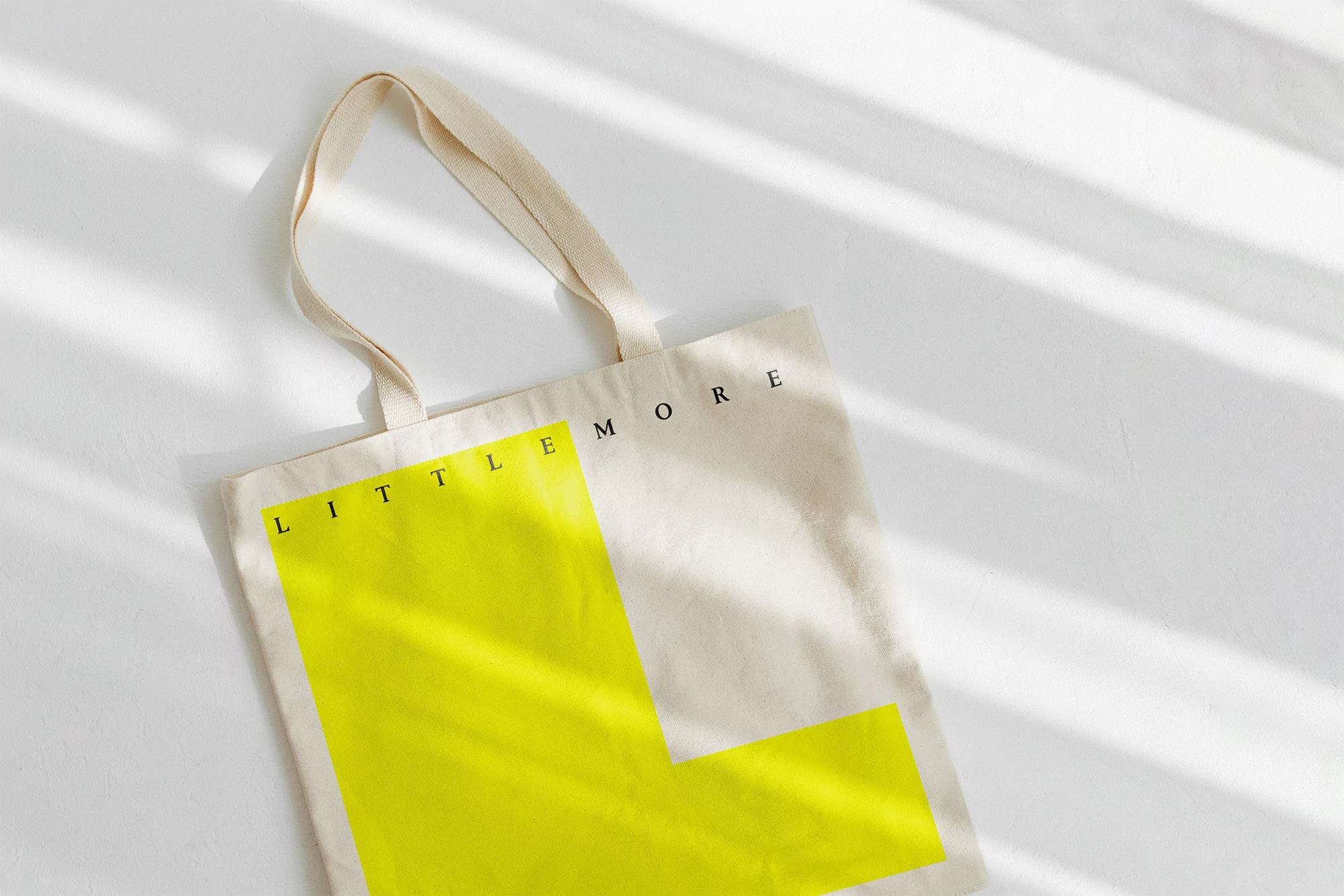 Beige tote bag with graphic L design in bright yellow.  Simple, modern advertisement on a white surface with light and shadow.