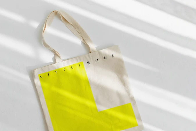 Beige tote bag with graphic L design in bright yellow.  Simple, modern advertisement on a white surface with light and shadow.