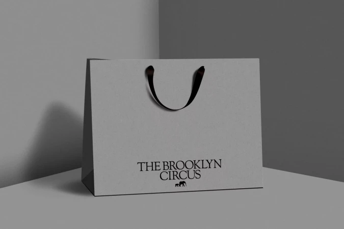 White shopping bag with black handles sits on a gray surface against a gray background featuring the text "The Brooklyn Circus."