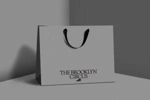 Grey paper shopping bag with black ribbon handles and black branded text set against a minimal corner backdrop.