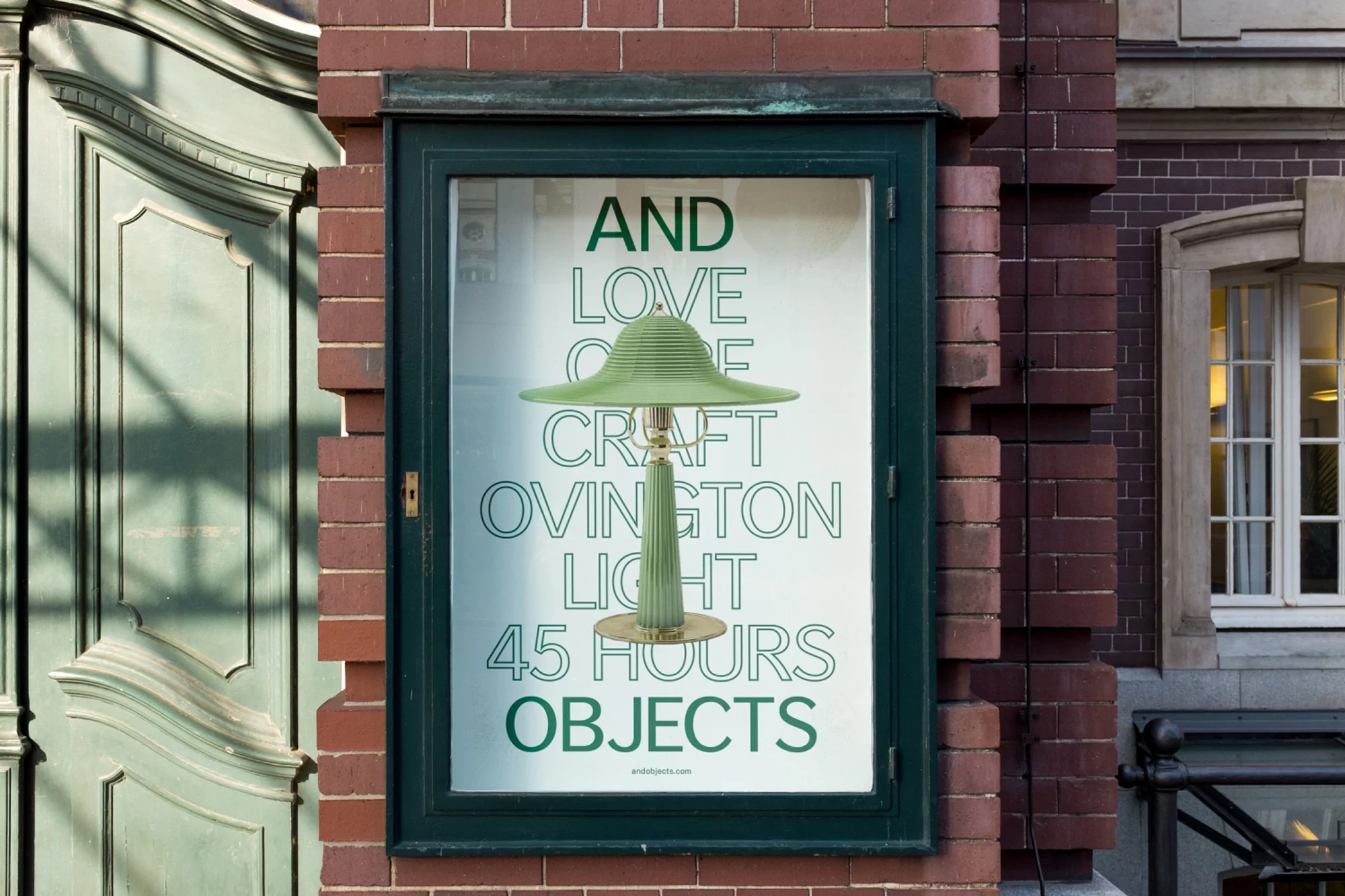 Window display of a green table lamp with a wide shade against a poster with green text, set in front of a brick wall and green door.