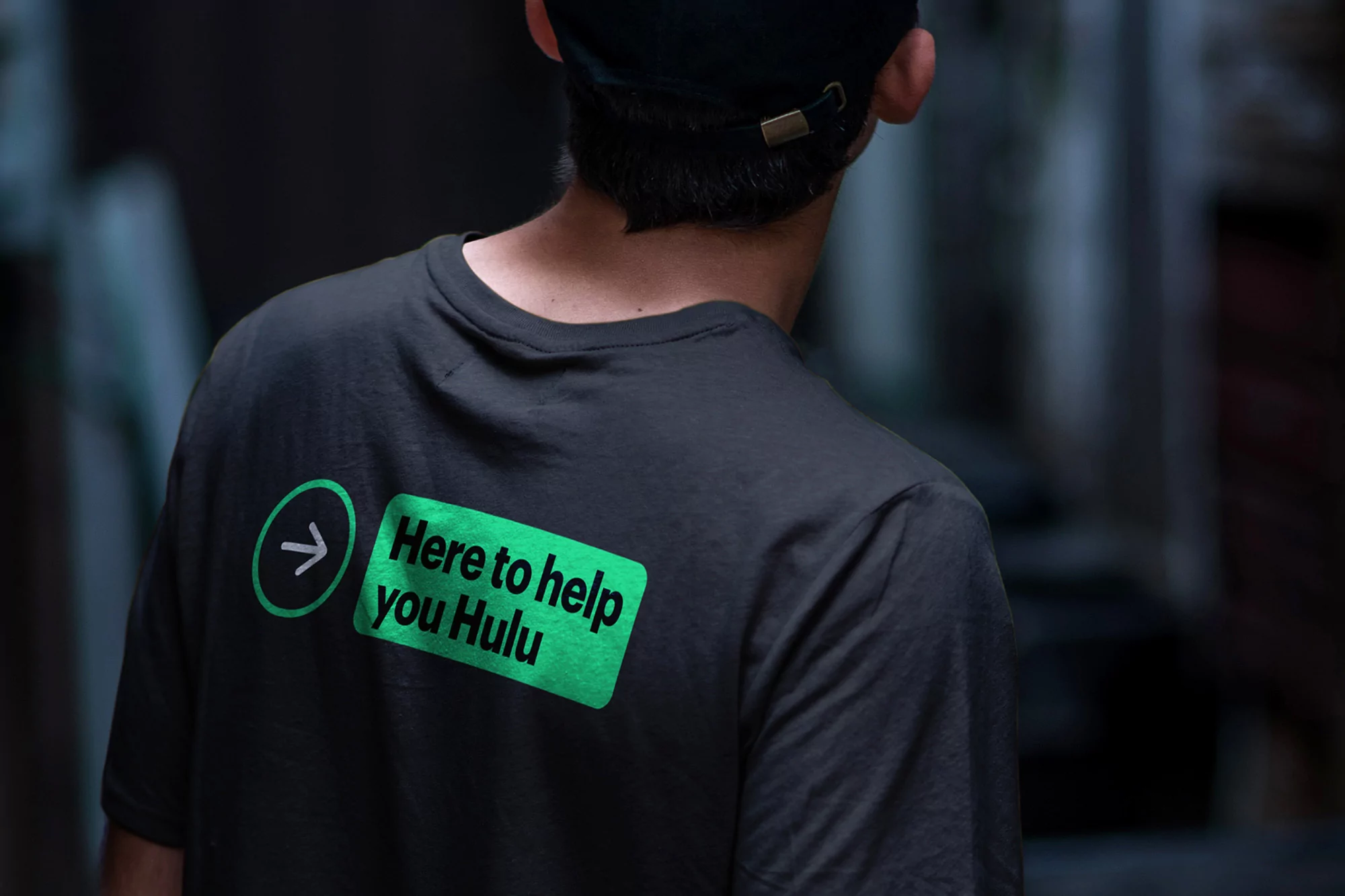 Person wearing a dark t-shirt with a Hulu logo and text on the back, viewed from behind in a blurred, neutral indoor space.