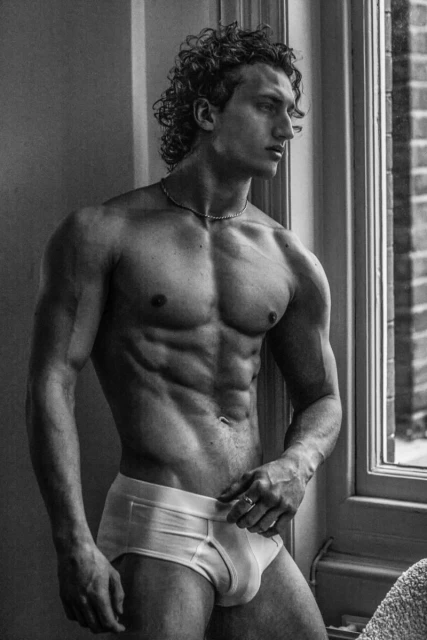 Black and white studio shot of a shirtless man with curly hair standing near a window, showcasing his muscular physique.