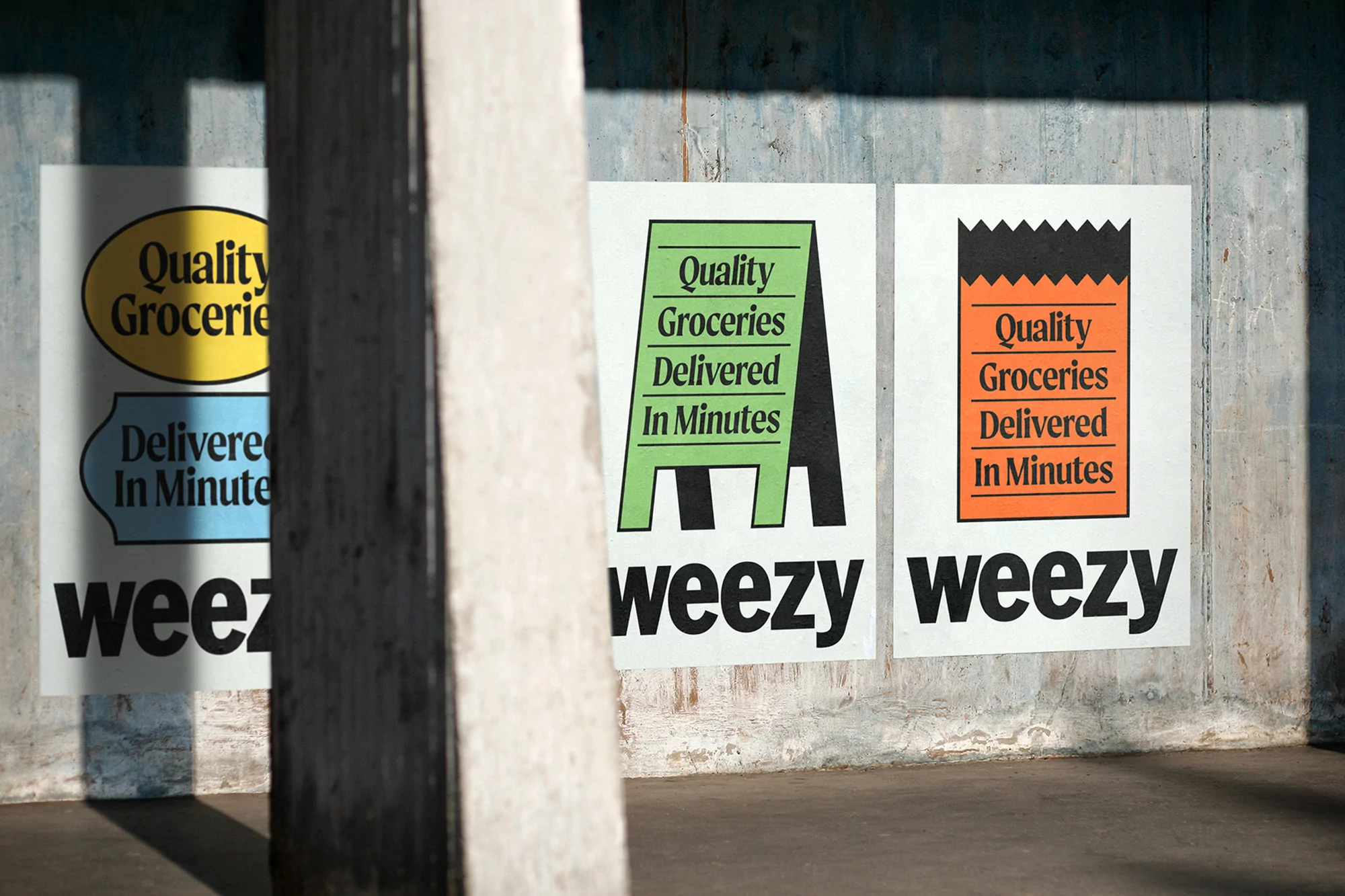 Three Weezly posters on a concrete wall advertise quality groceries and delivery in minutes, with a grey pillar partially obscuring one poster.