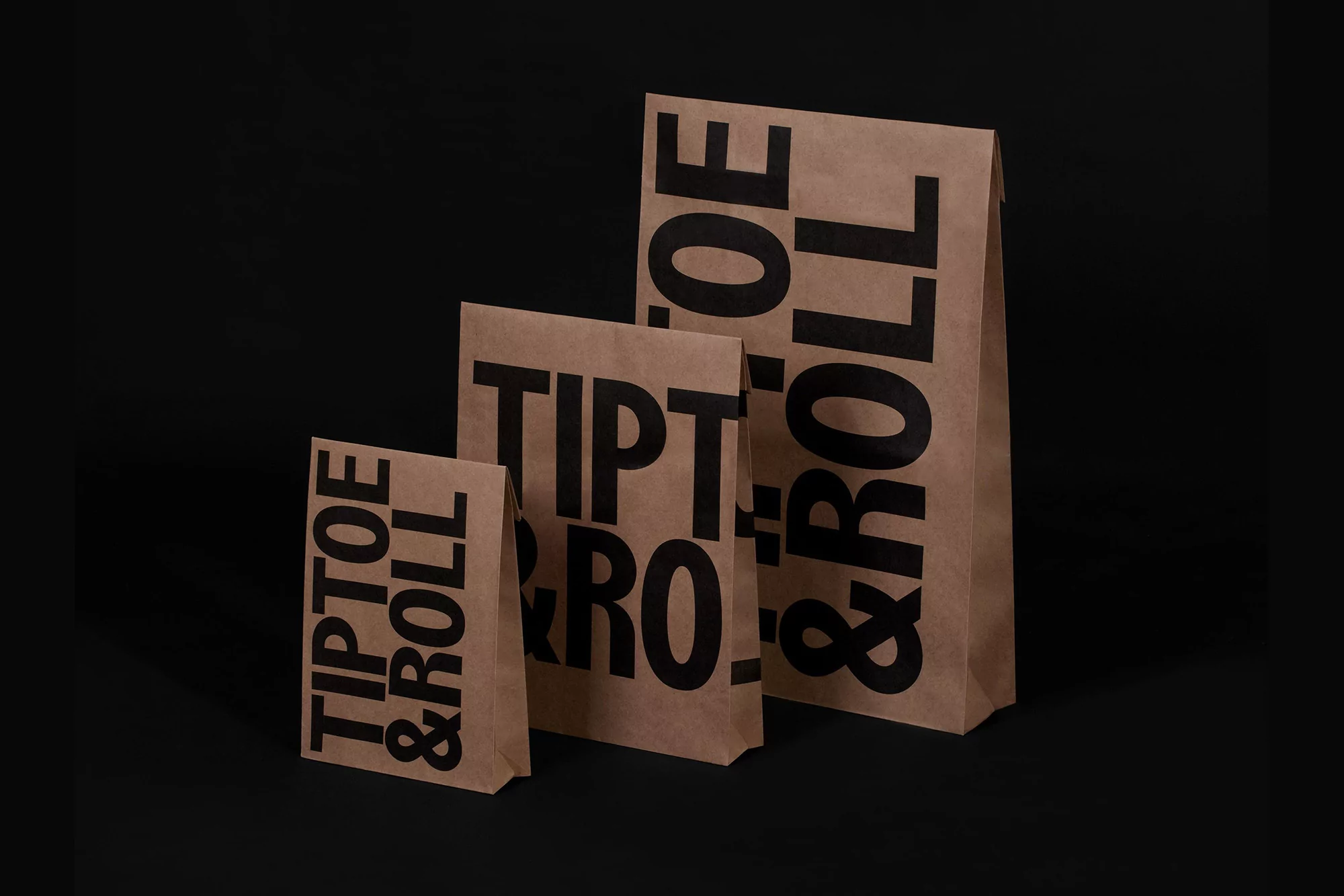 Three brown paper bags in different sizes, each printed with the words "TIPTOE & ROLL" in a bold, sans-serif black font, on a black background.
