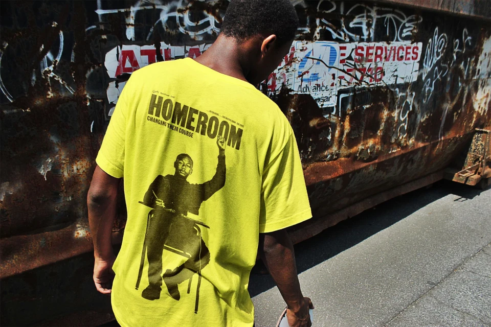 A person in a yellow t-shirt with a graphic of a student on it stands in front of a rusty metal dumpster in an outdoor urban setting.