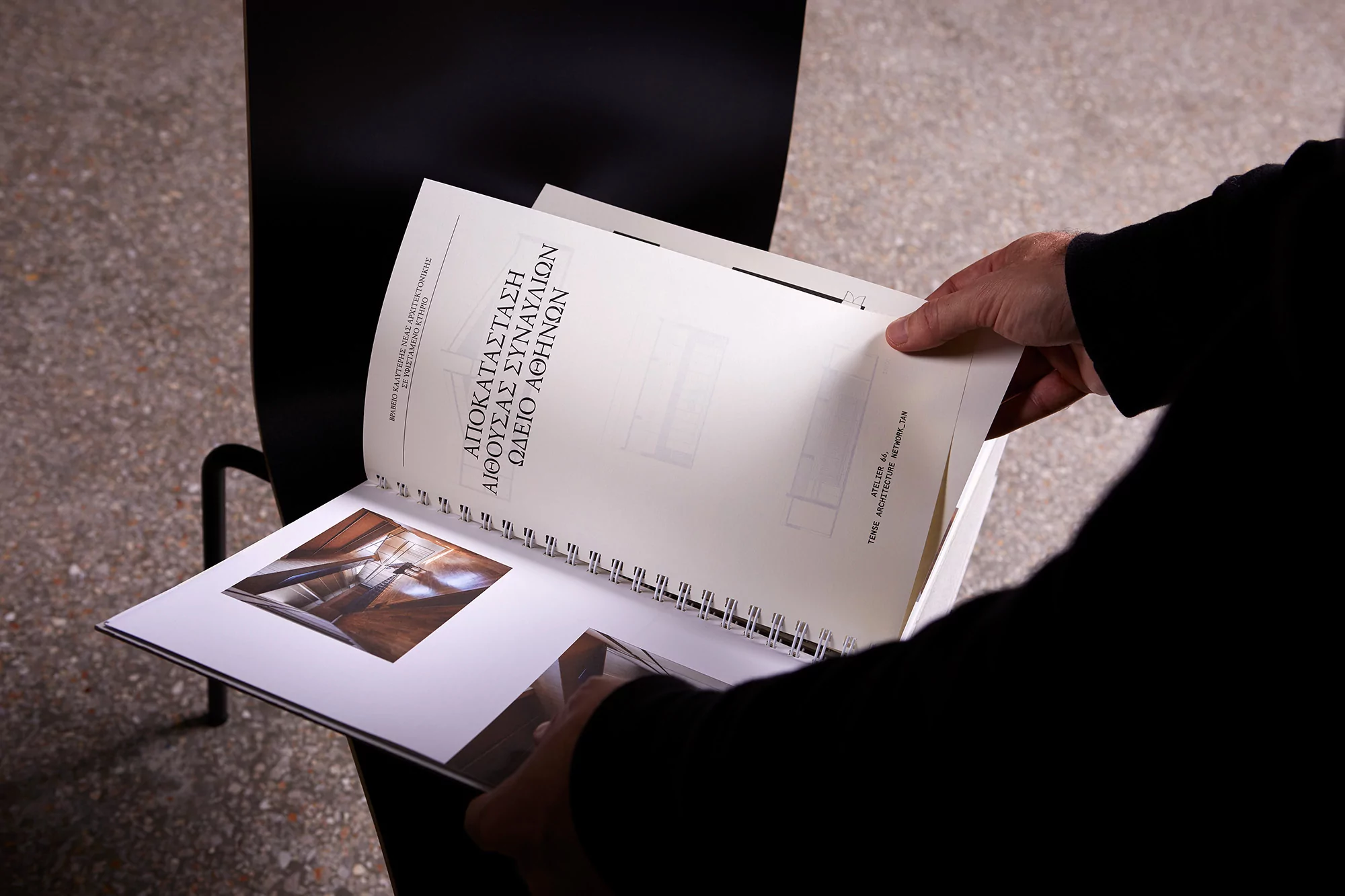 A person holds open a spiral-bound book, revealing architectural drawings and diagrams, with text in Greek. The setting is indoors.