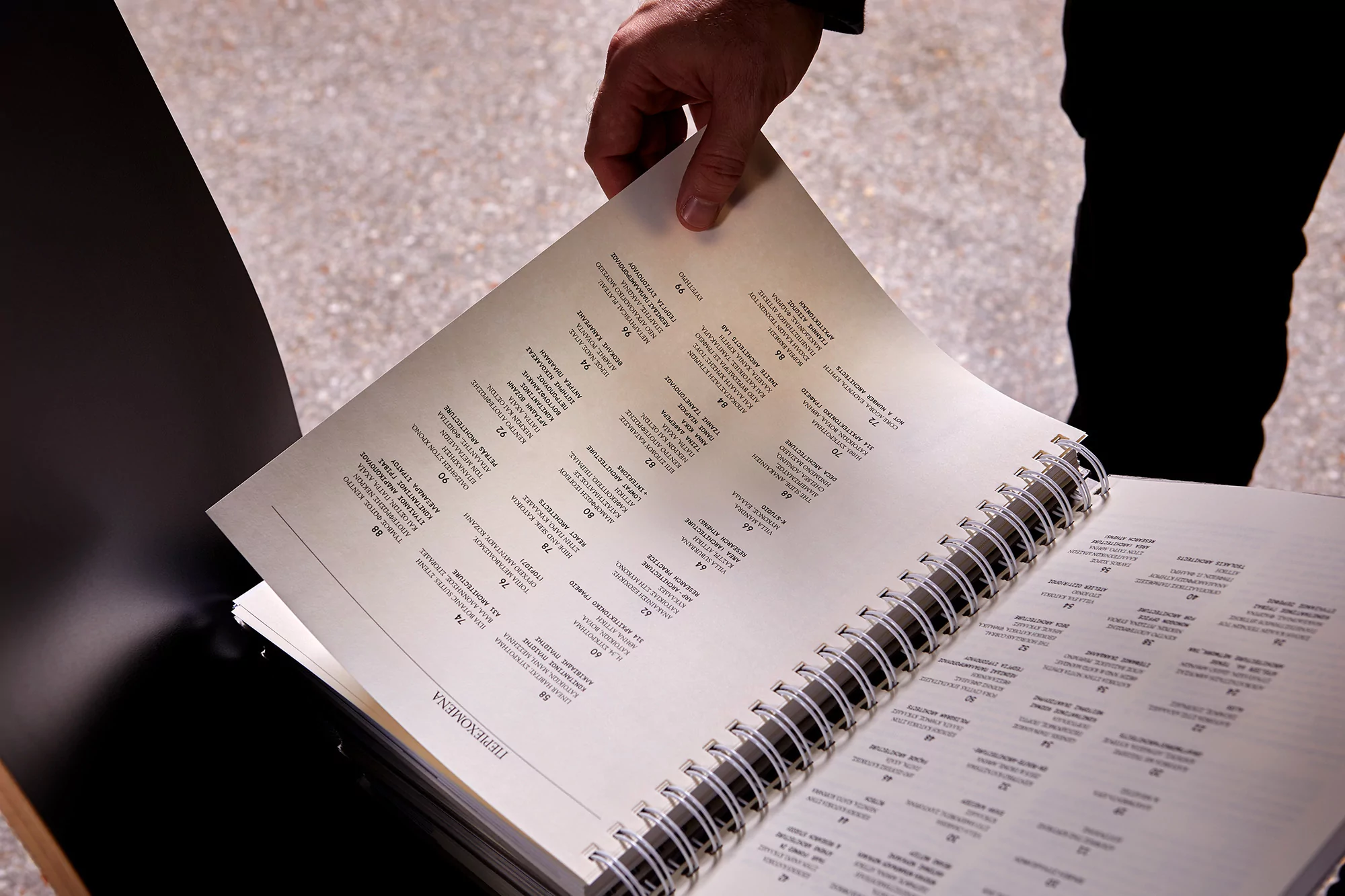 Person holds open a spiral-bound book with Greek text, offering a detailed look at its contents and binding, in a brightly lit scene.