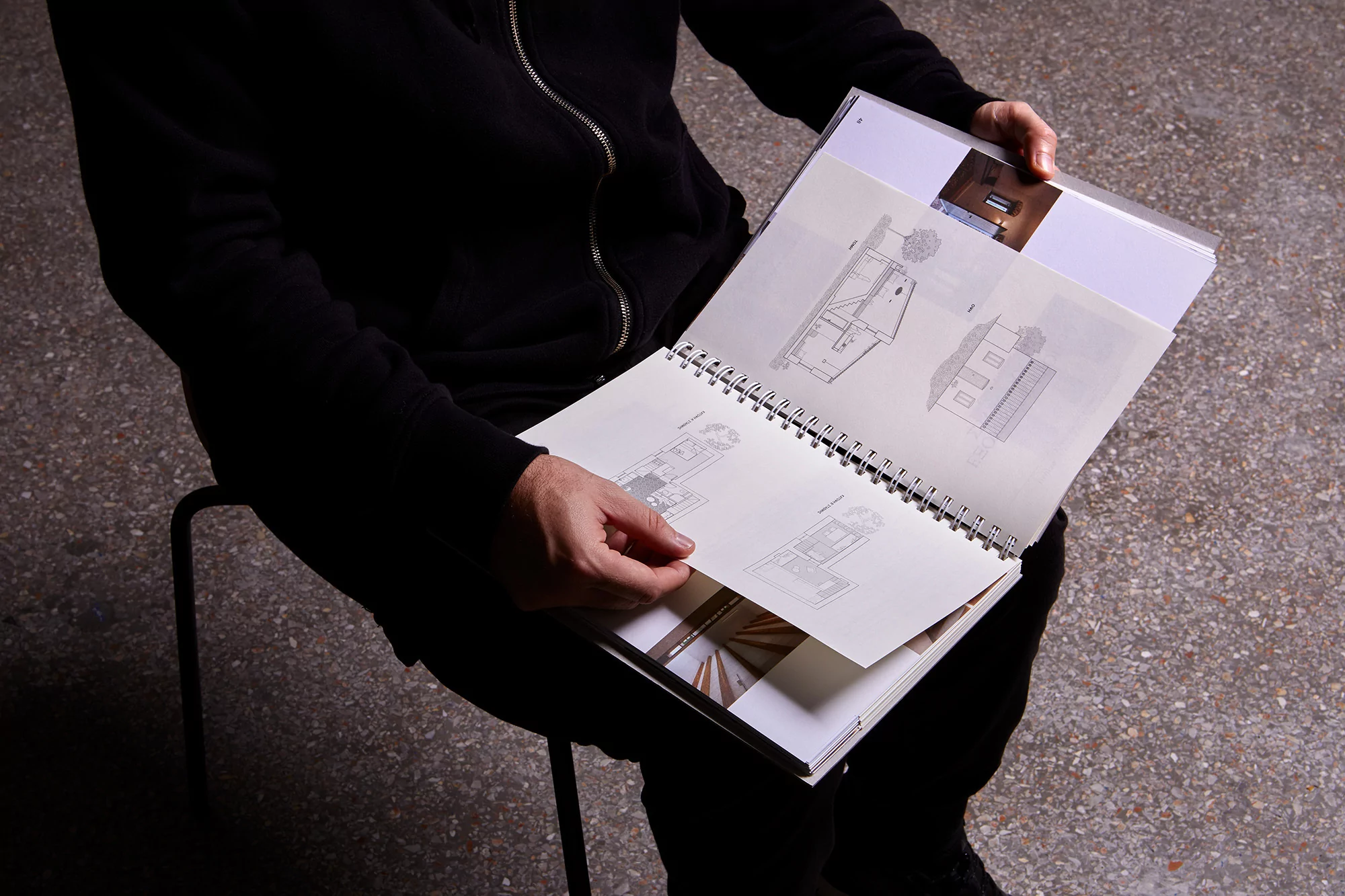 Person sitting on a chair, holding open a spiral-bound book displaying architectural sketches and interior photographs on the pages.