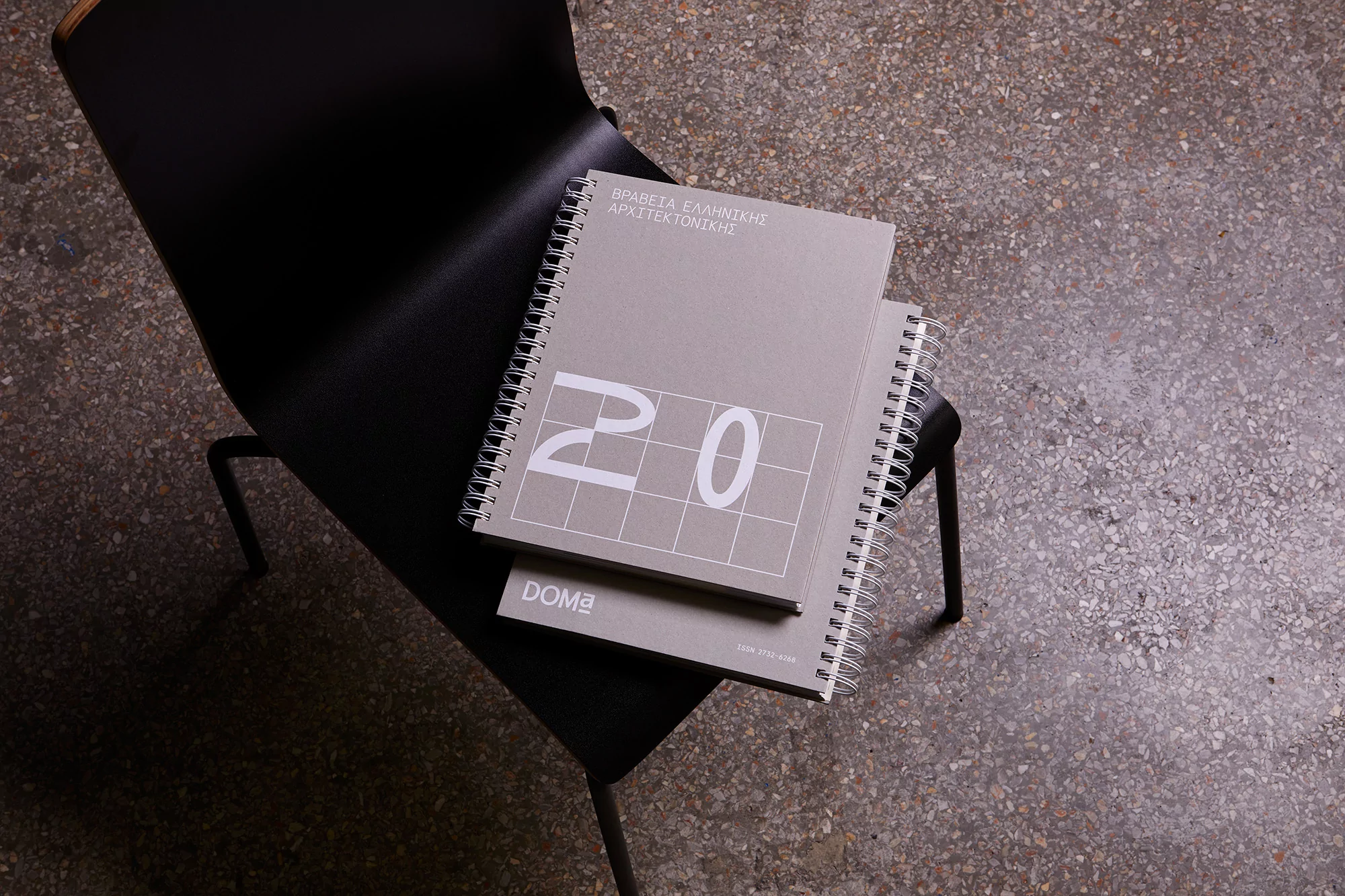 Two grey spiral notebooks with architectural designs on a black chair, photographed indoors with a neutral colour palette and concrete floors.