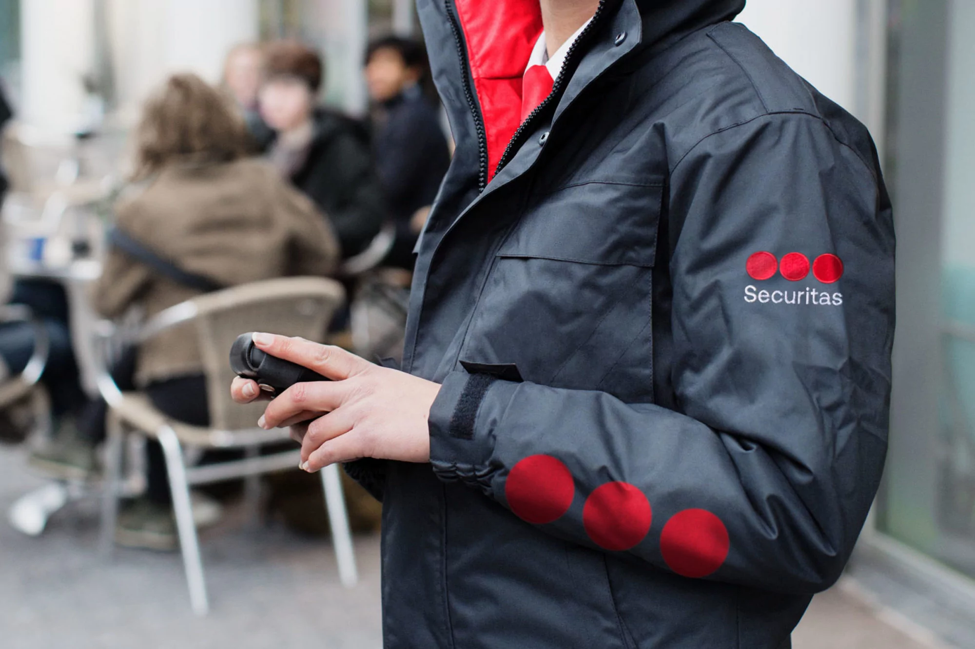 Person in a dark blue Securitas jacket, holding a black object, stands in an urban setting with people in the background.