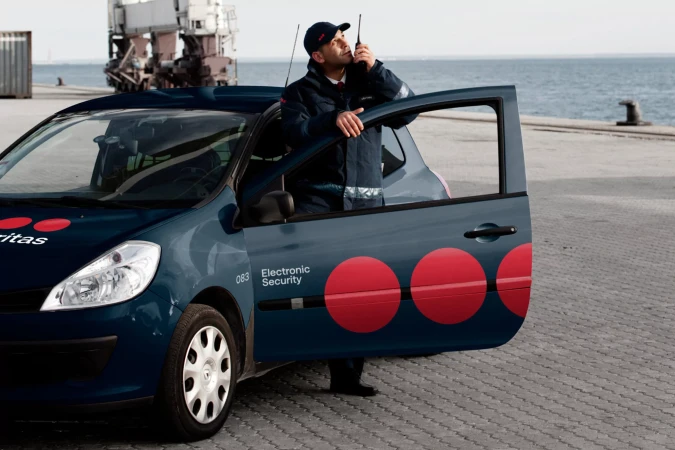 Security guard in uniform speaks into a walkie-talkie, standing next to a marked security vehicle with an open car door by the water.