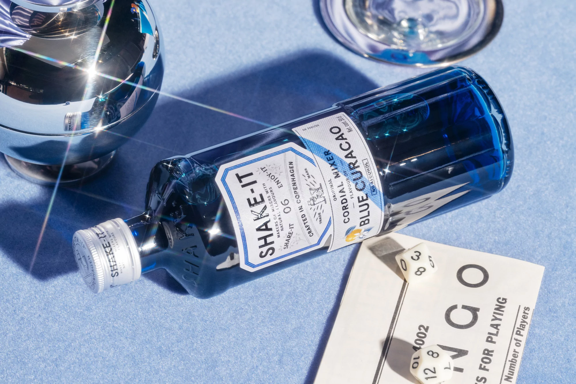 Studio shot of blue curaçao bottle on blue cloth with dice and bingo card; eye-level view with artificial light reflections.