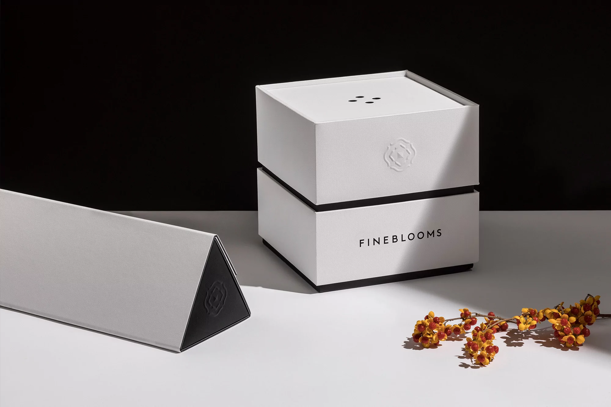 Elevated studio shot showcases two stacked white Fine Blooms boxes against a contrasting black backdrop and a sprig of berries.