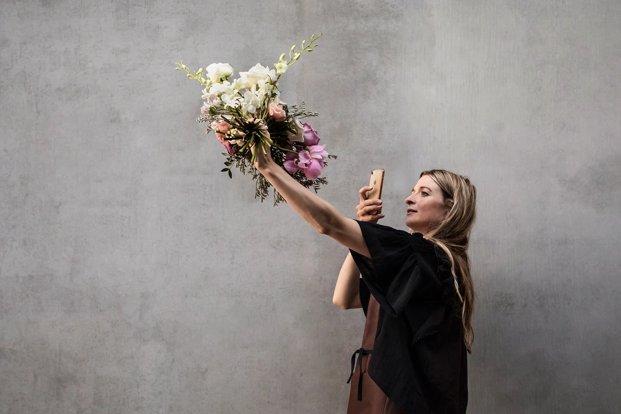 Woman in black top holds a bouquet of white, pink and purple flowers, taking a photo with her smartphone against a gray background.