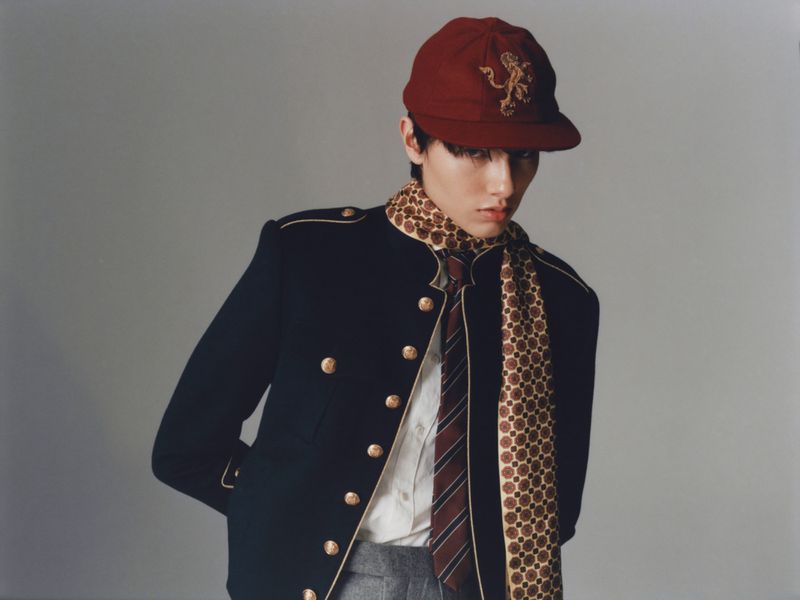 Portrait of a young adult in a dark navy jacket with gold buttons, maroon cap with gold emblem, and patterned scarf on a grey background.