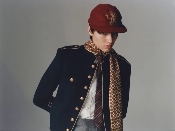 Portrait of a young adult in a dark navy jacket with gold buttons, maroon cap with gold emblem, and patterned scarf on a grey background.