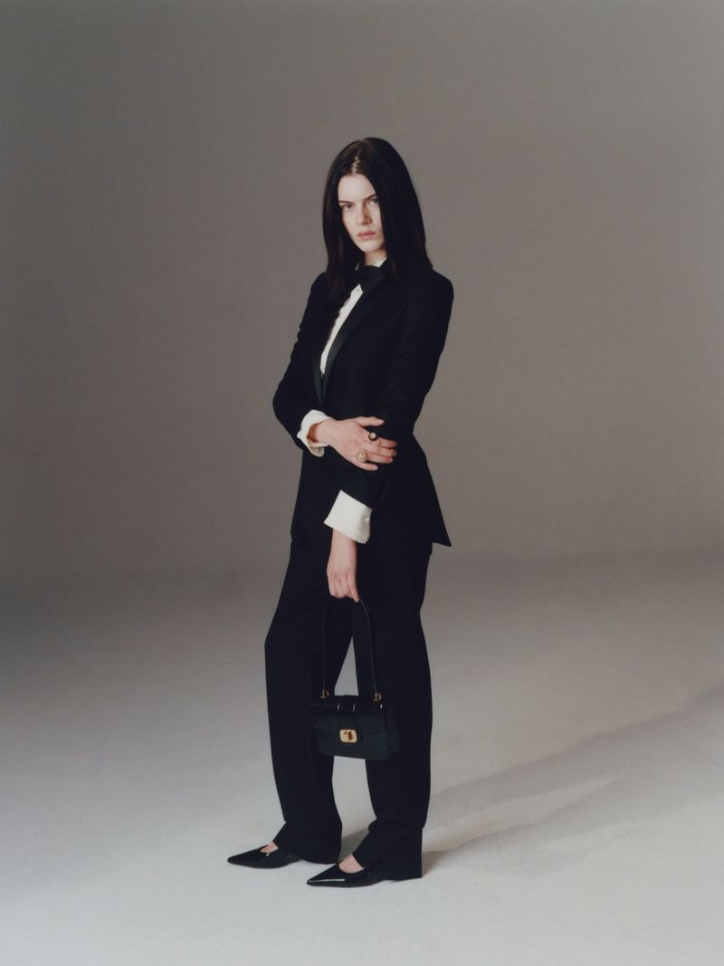 Elegant woman in a black suit with a handbag, captured in a studio setting with soft lighting, showcasing a blend of classic and modern fashion.