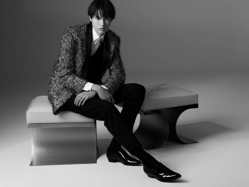 Black and white portrait of a young man in a shimmering jacket sitting on a modern bench in a neutral-toned photo studio.