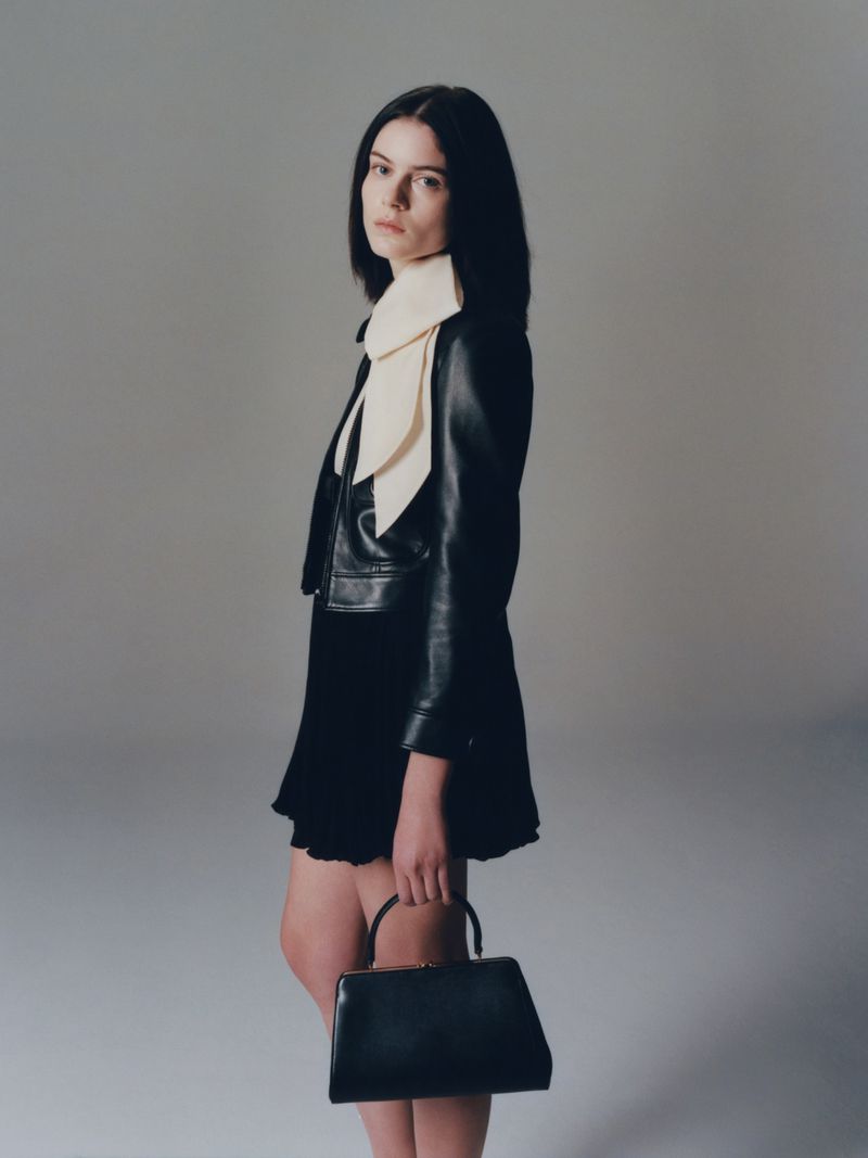 A brunette woman poses indoors wearing a black jacket, black dress, and carrying a black handbag. She faces the camera with a neutral expression.