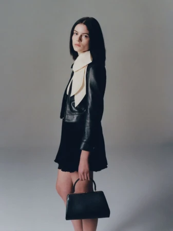 A brunette woman poses indoors wearing a black jacket, black dress, and carrying a black handbag. She faces the camera with a neutral expression.