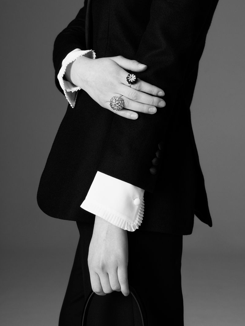 Close-up black and white image of a person wearing a black suit, white ruffled shirt cuffs, and rings, holding a hat.