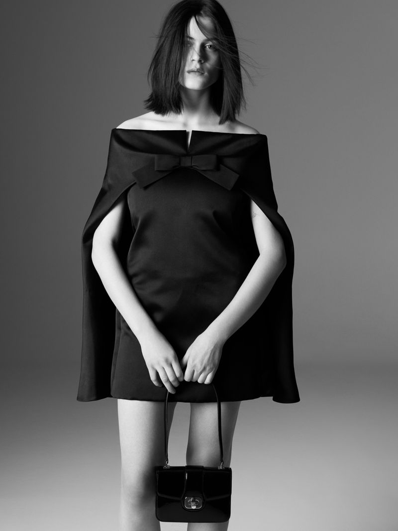 Monochrome studio portrait of a woman wearing a black dress with a bow and holding a black purse against a grey background.