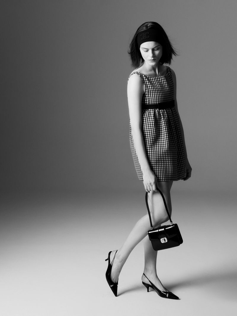 A young woman in a patterned mini dress poses in a studio with a purse and heels. Black and white portrait showcases fashion.