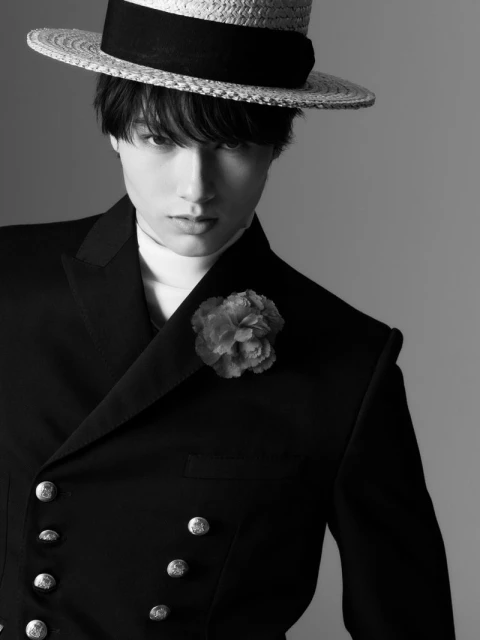 Black and white studio portrait of a young adult man in a double-breasted suit, straw hat, and flower lapel against a grey backdrop.