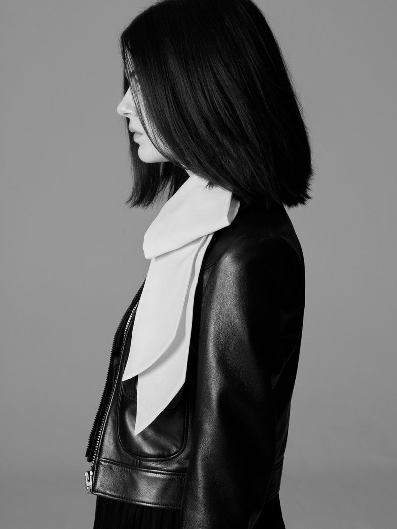 Profile shot of a woman with short dark hair wearing a black leather jacket and white scarf against a gray studio backdrop.