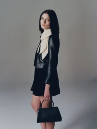 Model wearing a black leather jacket and carrying a handbag against a neutral grey studio background