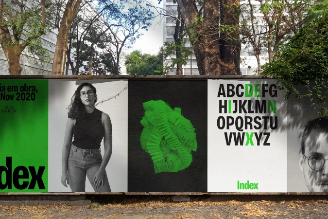 Outdoor poster exhibit with portrait of woman, abstract green shape, and alphabet art. Urban setting with trees and building visible.