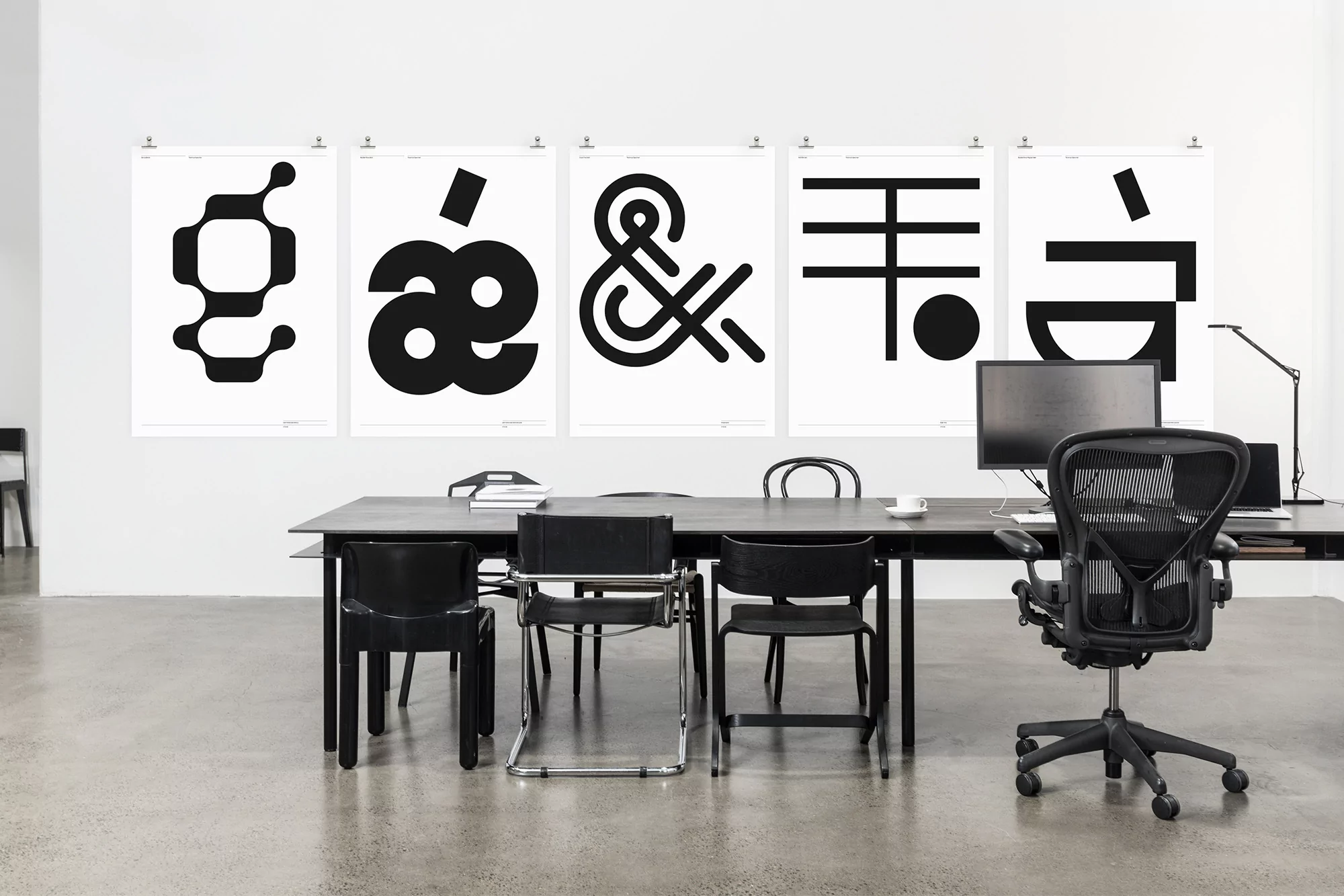 Modern workspace featuring a black desk, chairs, and computer, with five graphic black and white posters hanging on the white wall.