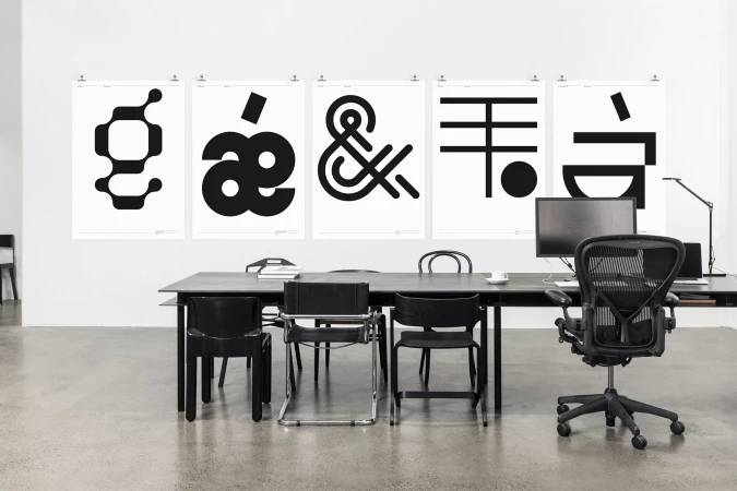 Modern workspace featuring a black desk, chairs, and computer, with five graphic black and white posters hanging on the white wall.