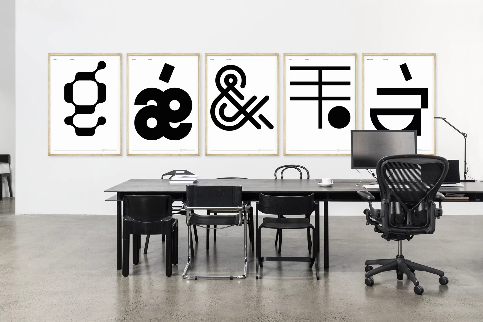 Office space featuring black desk, black chairs, a computer, and framed typographic prints in a minimalist, modern interior setting.