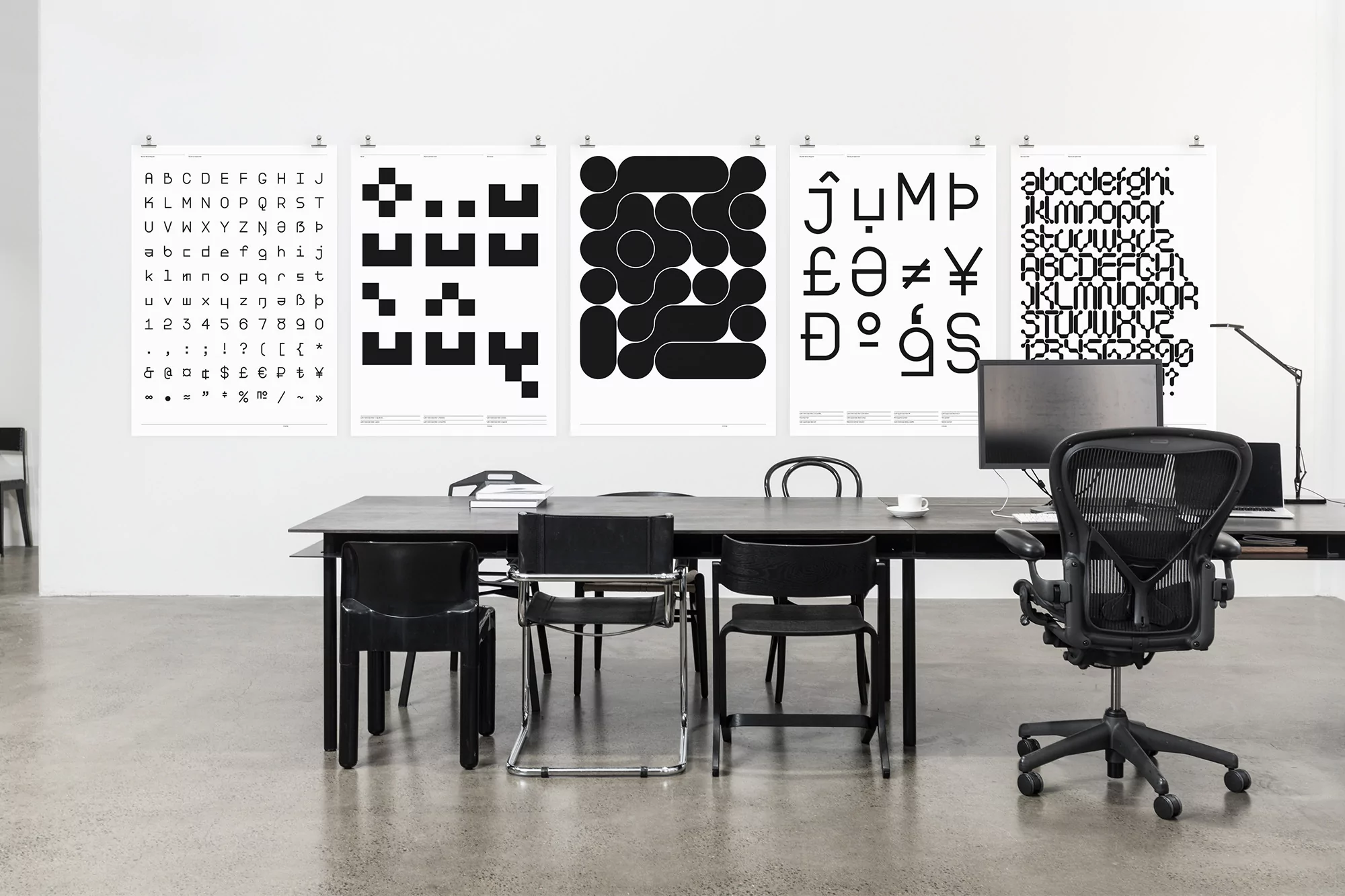 Office interior with five posters hanging on the wall, featuring bold black and white geometric designs and typography; black desk and chairs.