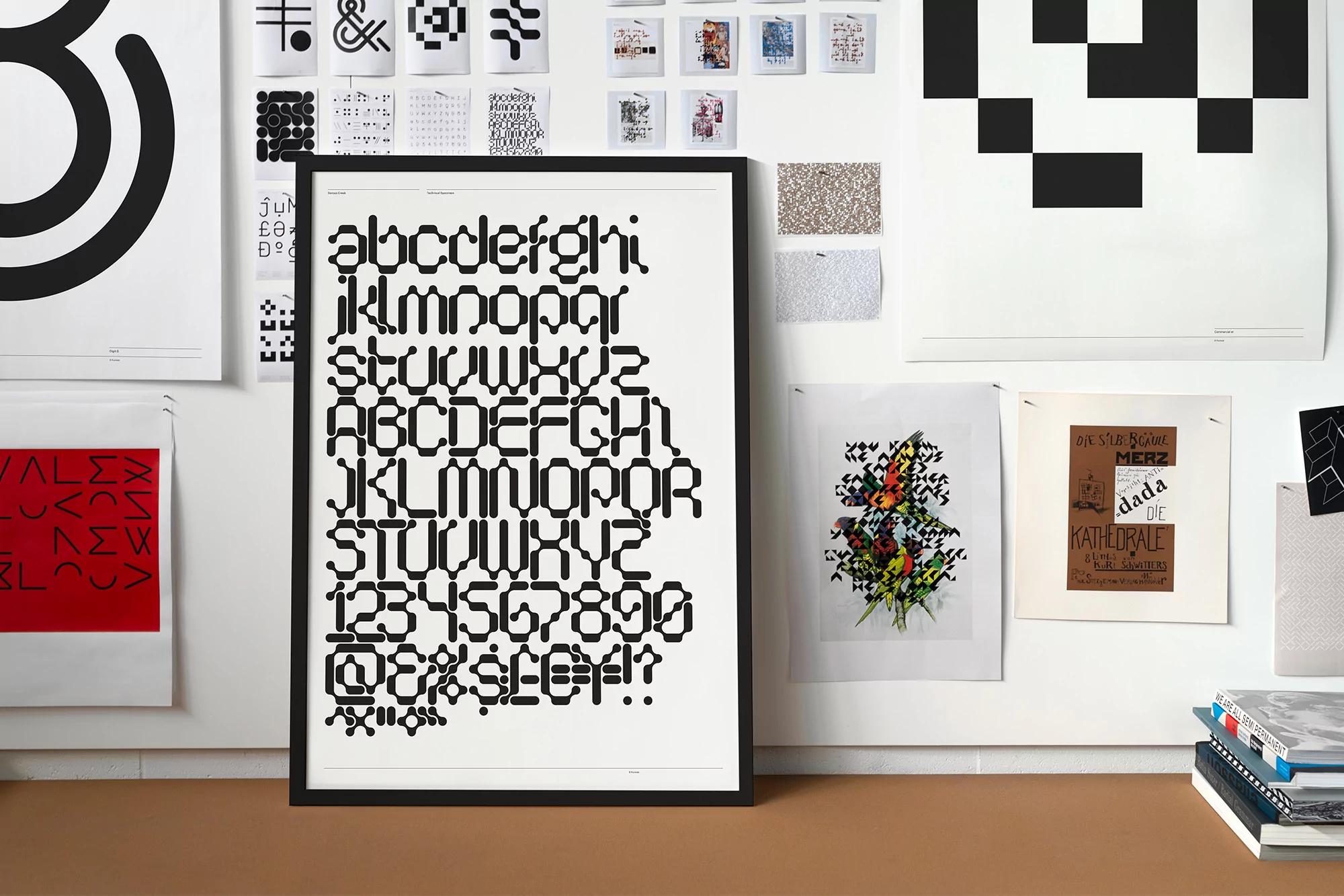 Framed poster of black block-style alphabet characters on a white wall, surrounded by other prints, typography, and design ephemera.
