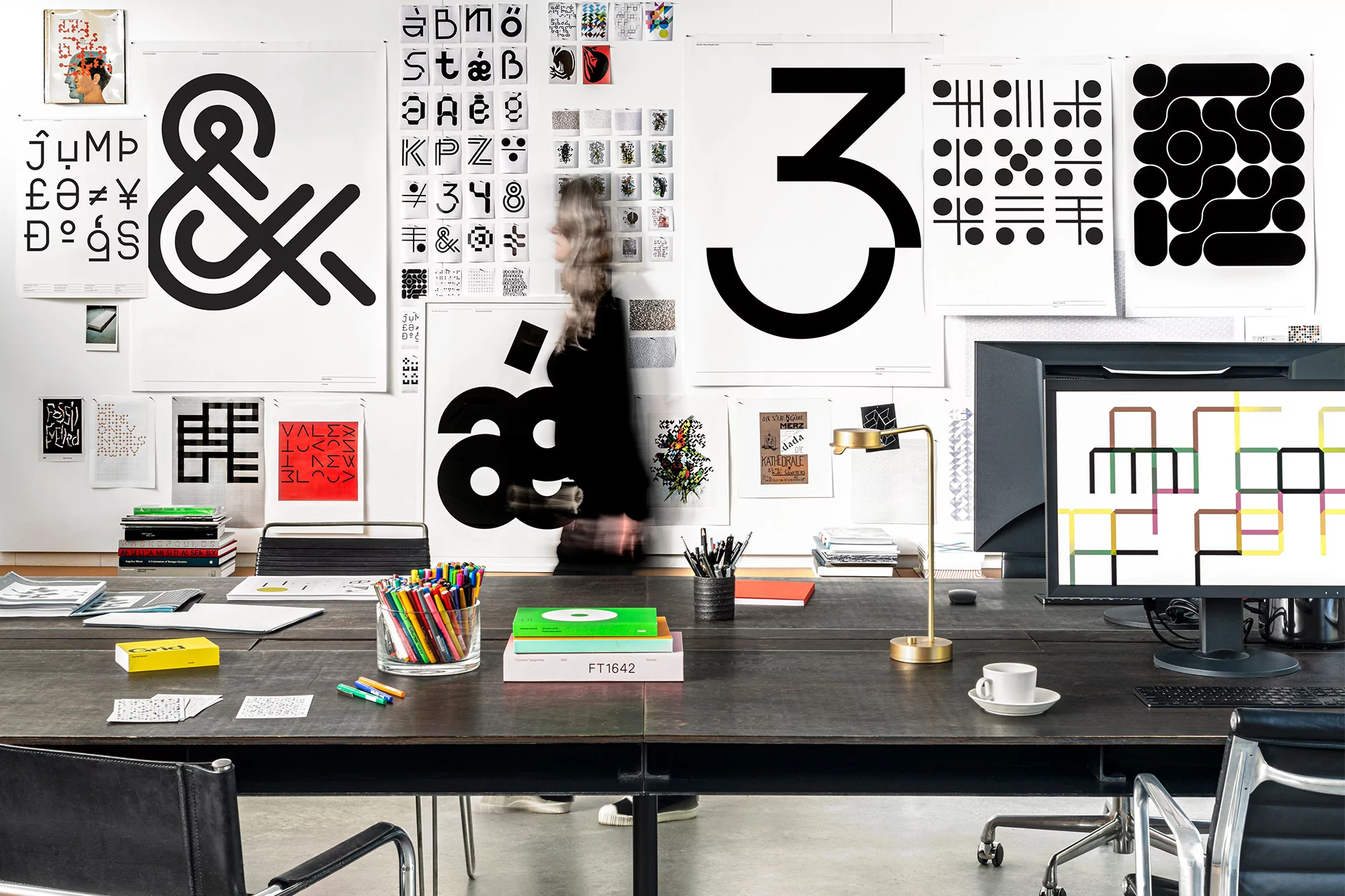 Workspace with a large wooden desk, various posters with graphic designs, a computer, and office supplies in a brightly lit interior.