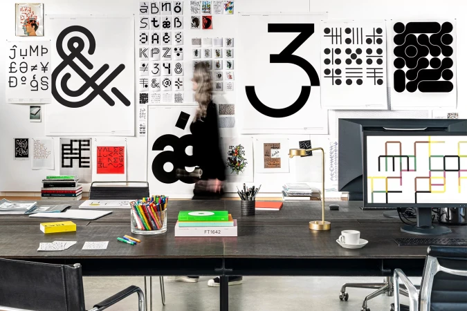 Workspace with a large wooden desk, various posters with graphic designs, a computer, and office supplies in a brightly lit interior.