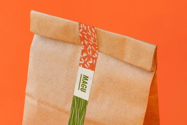 Brown paper takeaway bag sealed with patterned tape featuring the text MAGU against an orange background