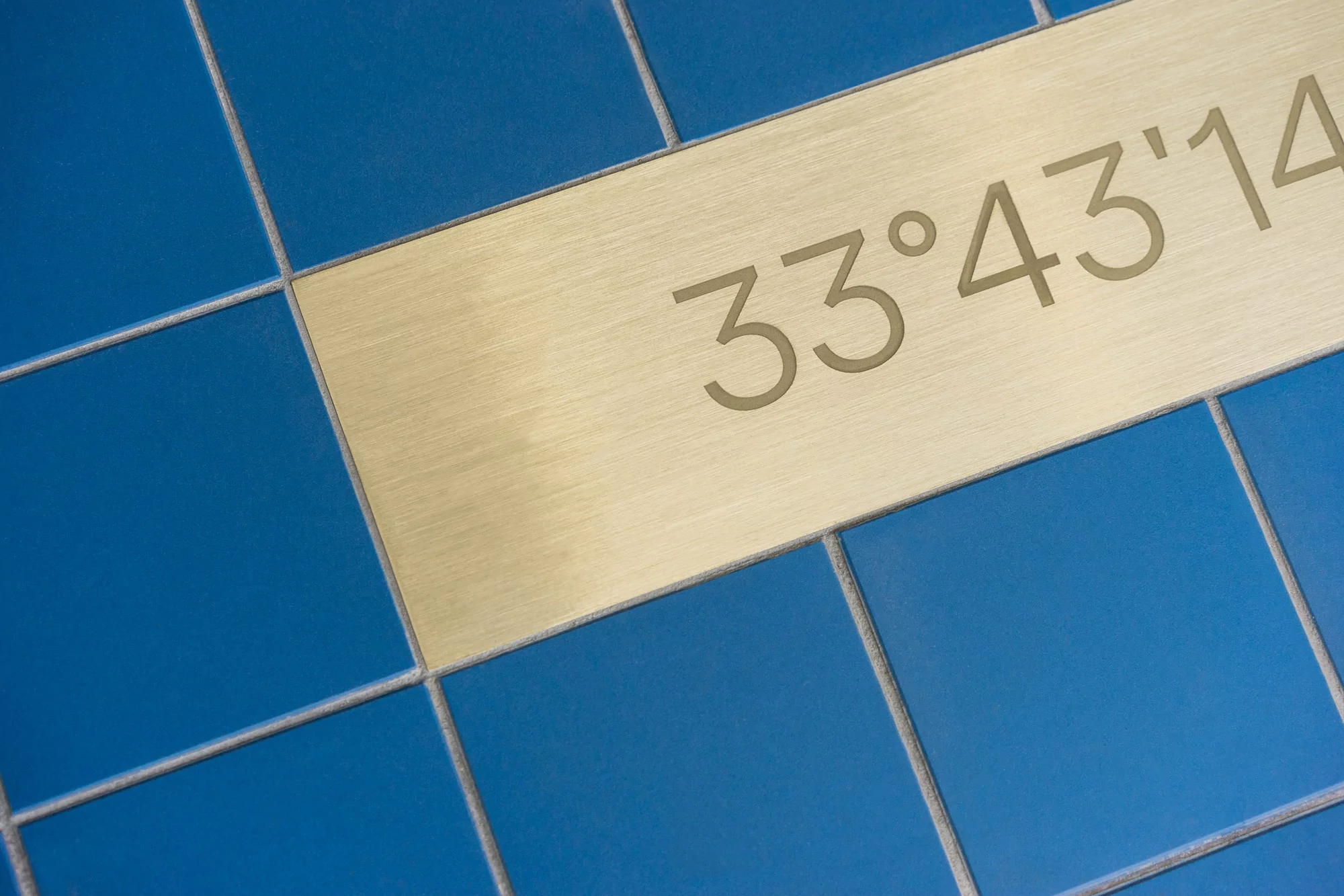 Close-up of blue ceramic tiles with a gold metal strip displaying the text '33°43'14,' creating a geometric pattern.